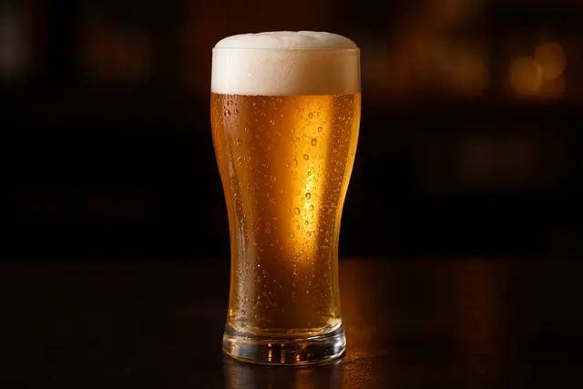 Tall pilsner glass of pale golden lager with a white head on a dark bar top